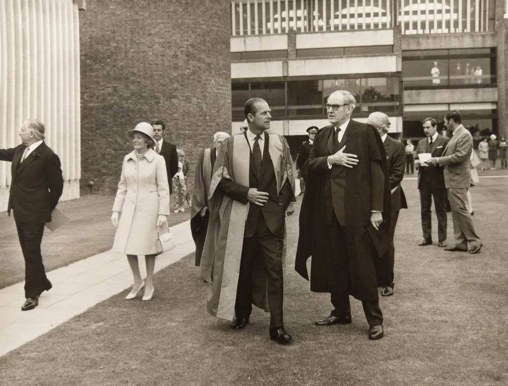 A group of people walks on a grassy area near a modern building. Two men in academic gowns are at the forefront, engaged in conversation. A woman in a coat and hat follows them, with other individuals visible in the background. The scene is in black and white.