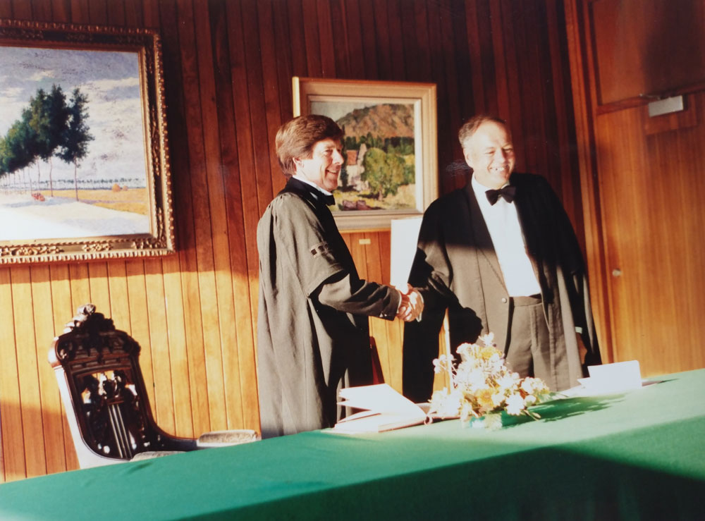 Two people in formal attire shake hands across a table with a green cloth. A bouquet of flowers and papers are on the table. The room has wooden walls and is adorned with framed paintings. Warm light illuminates the scene.