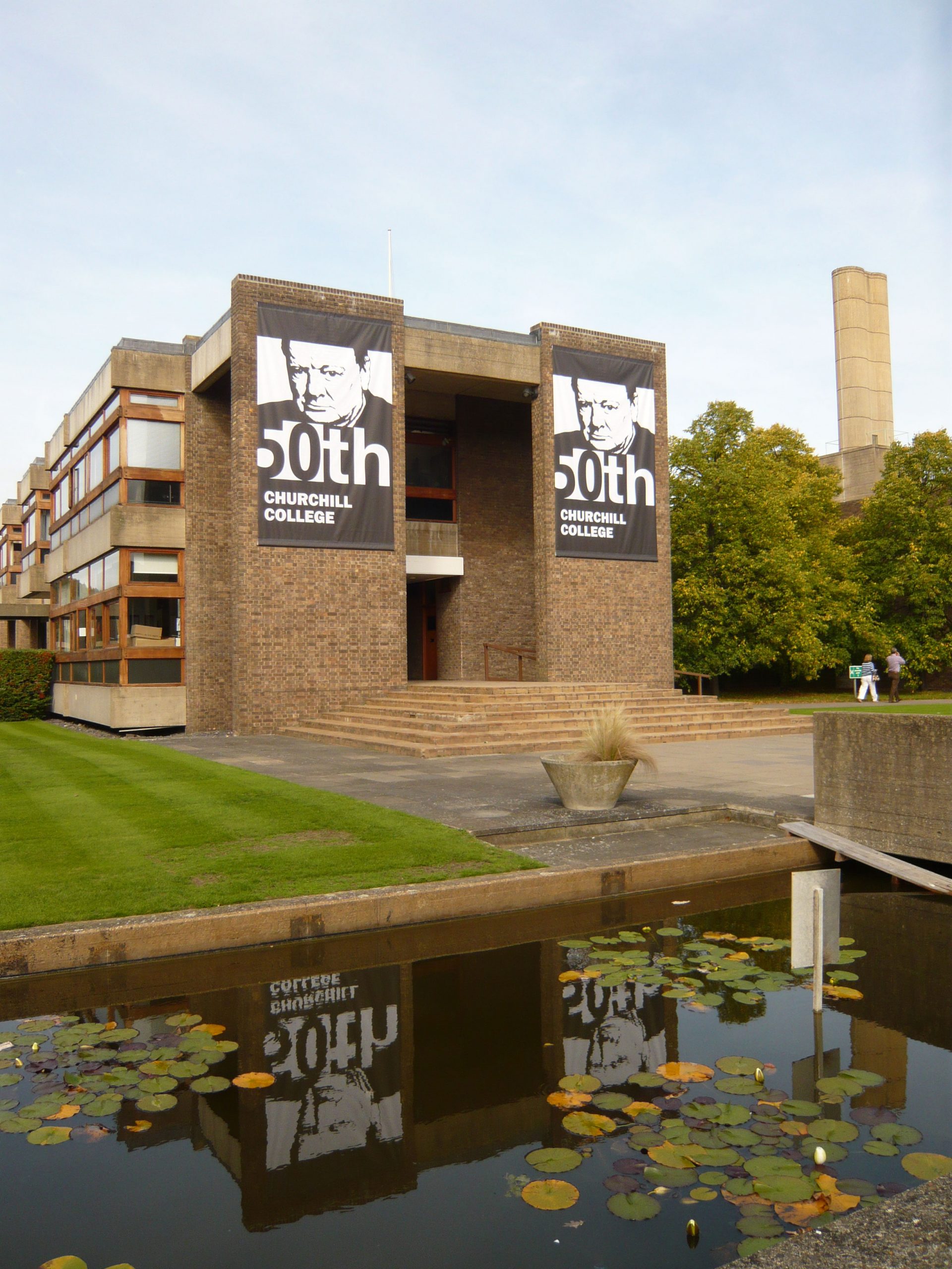 A brick building with large banners featuring 50th anniversary of Churchill College. A pathway leads to the entrance, flanked by green grass. In the foreground, a pond with lily pads and a sculpture. A chimney-like structure and trees are visible in the background.