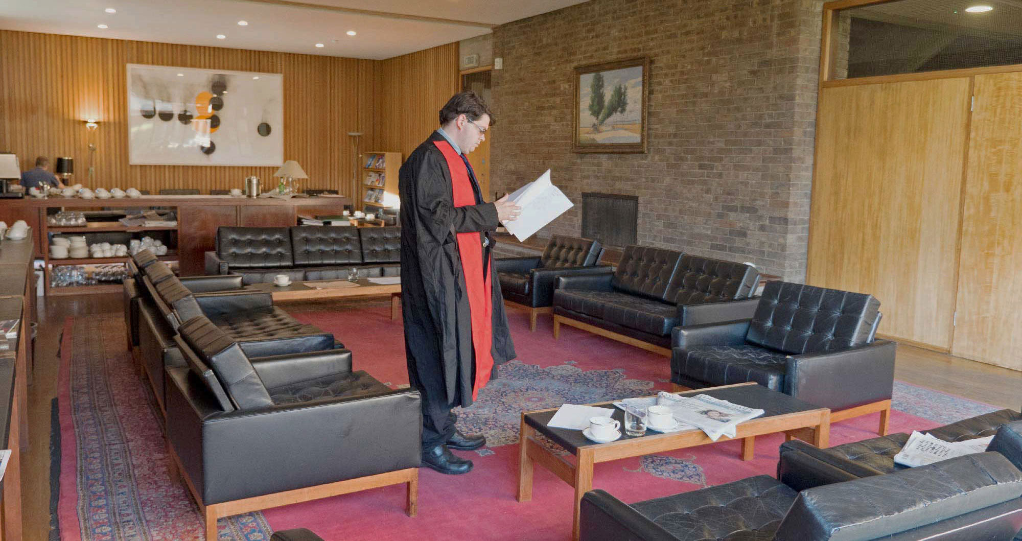 A person wearing a robe with a red scarf stands in a mid-century modern room reading a document. The room features dark leather chairs, wooden paneling, a brick wall, art, and a table with cups and saucers. Newspapers are on another table.