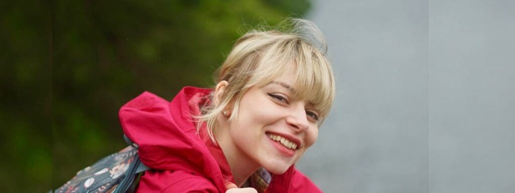 A person with blonde hair smiles while wearing a bright red jacket and carrying a patterned backpack. The background is a mix of greenery and a gray wall.