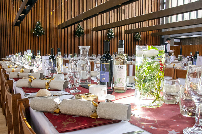A festive banquet table set for a gathering, featuring wine bottles, glassware, candles, and Christmas crackers. The room is warmly lit with wooden décor and holiday decorations, including greenery and lights, enhancing the celebratory atmosphere.