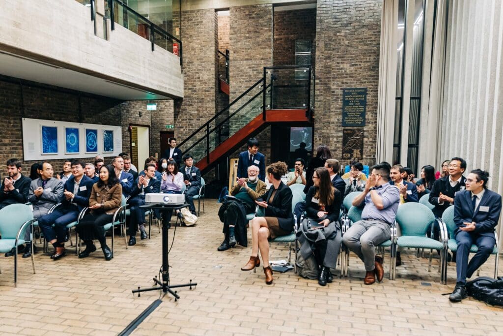 A group of people sitting on chairs in a brick-walled room with framed artwork. A projector is in the foreground. Some individuals are clapping, while others are watching intently. A staircase is visible in the background.