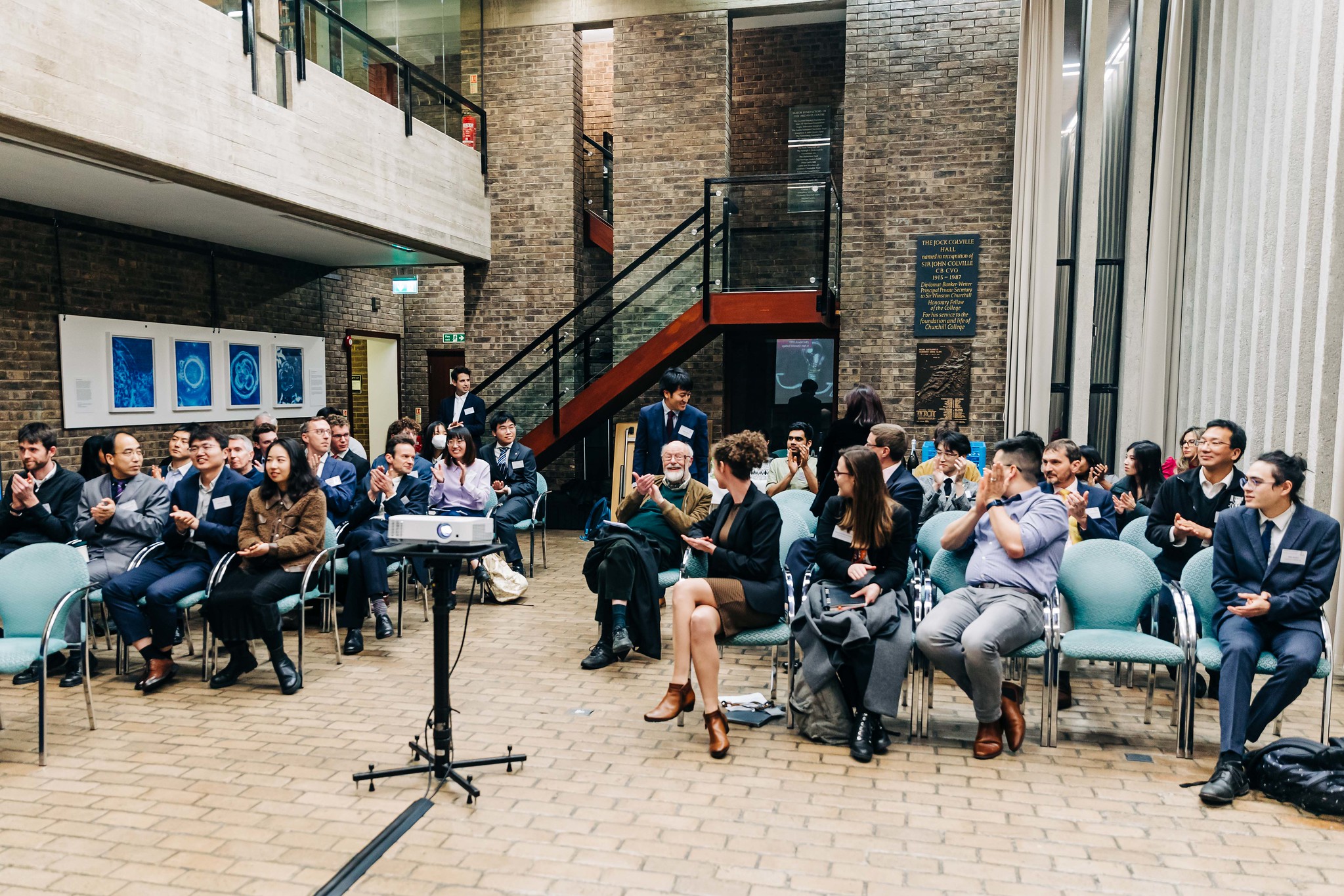 A group of people sitting on chairs in a brick-walled room with framed artwork. A projector is in the foreground. Some individuals are clapping, while others are watching intently. A staircase is visible in the background.