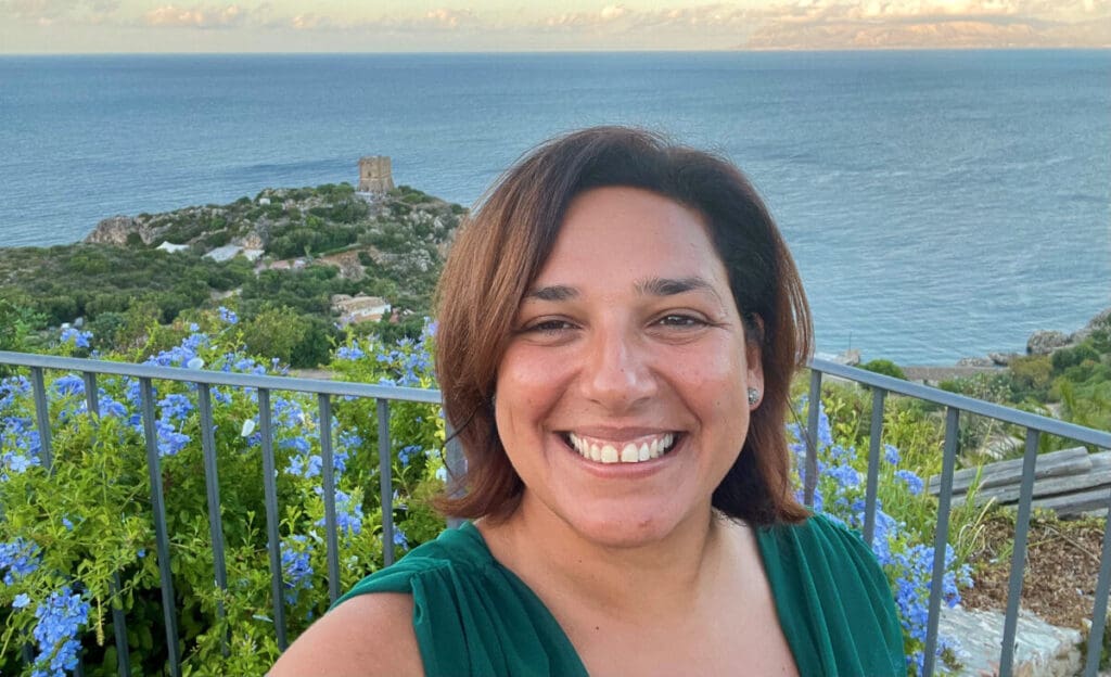 Woman smiling in a selfie with the ocean and a scenic coastline in the background. She stands near blue flowers, and theres a historic tower visible in the distance on a hilltop. The sky is partly cloudy.