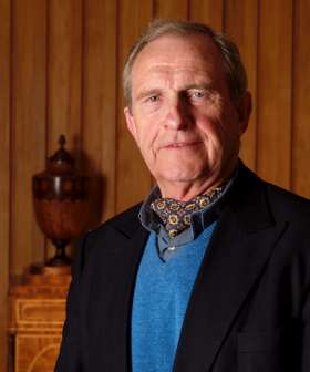 A man with short gray hair wearing a blue sweater, patterned scarf, and black jacket is standing in front of a wooden wall with an ornate vase in the background.
