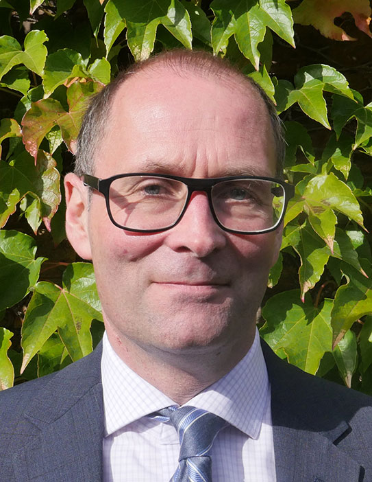 A man wearing glasses, a white checkered shirt, a blue tie, and a dark suit is standing in front of a backdrop of green leaves. He is facing the camera with a neutral expression.
