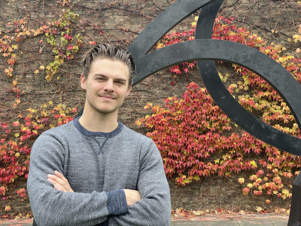 A person with short hair and a striped sweater stands with arms crossed in front of a large black sculpture. The background features a wall covered in vibrant red and orange ivy leaves.
