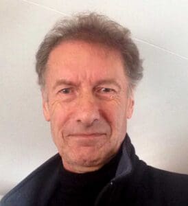 An older man with gray hair and a neutral expression is looking at the camera. He is wearing a dark jacket over a black top. The background is plain and light-colored.