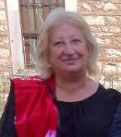 A woman with white hair, wearing a black top and a red scarf draped over her shoulder, stands in front of a stone wall with a window.