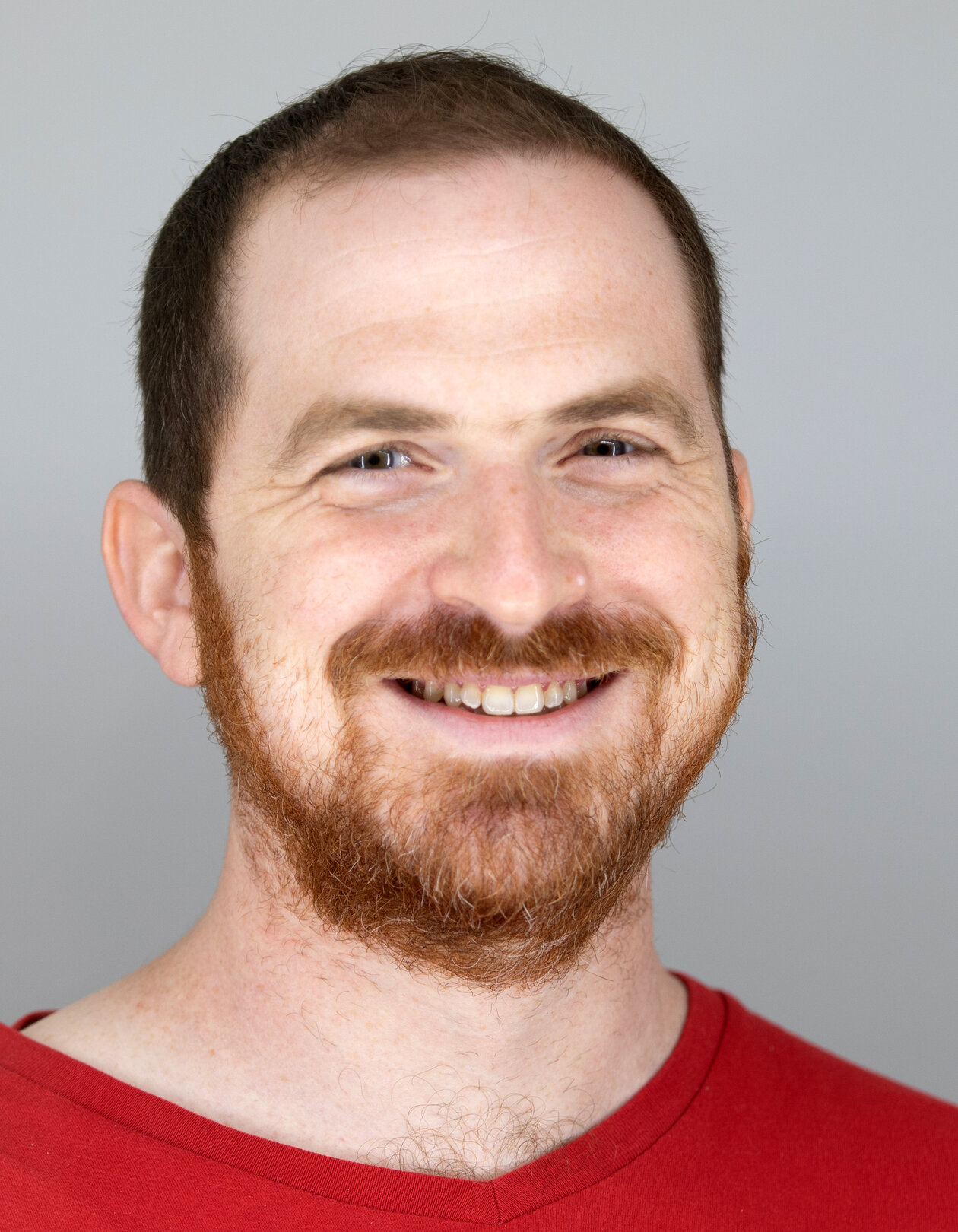 A man with a reddish-brown beard and short hair smiles warmly. He is wearing a red shirt, and the background is plain light gray.
