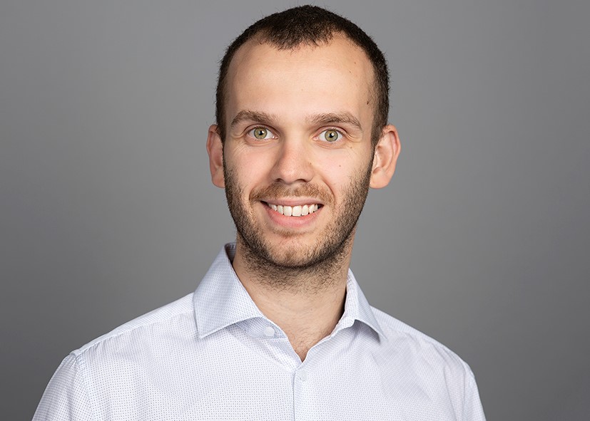 Portrait of a smiling man with short hair and a beard, wearing a light-colored button-up shirt against a gray background.