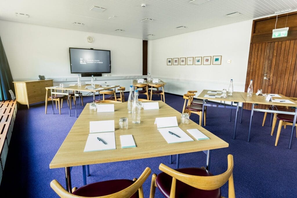 A well-lit conference room featuring three wooden tables arranged in a U-shape, each with notebooks, pens, water bottles, and glasses. A large screen on the wall displays a presentation. Framed art decorates the white walls, and a wooden door is on the right.