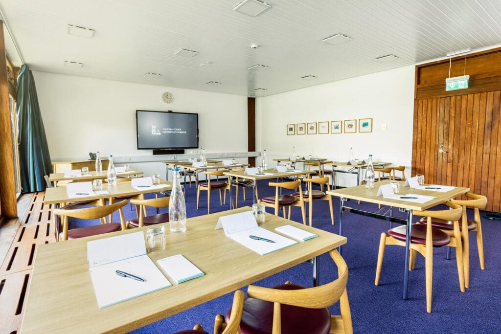 A bright conference room with several wooden tables and chairs facing a large screen displaying text. Each table has a notebook, pen, and water bottle. The walls are decorated with a few framed pictures, and there is a blue carpet on the floor.
