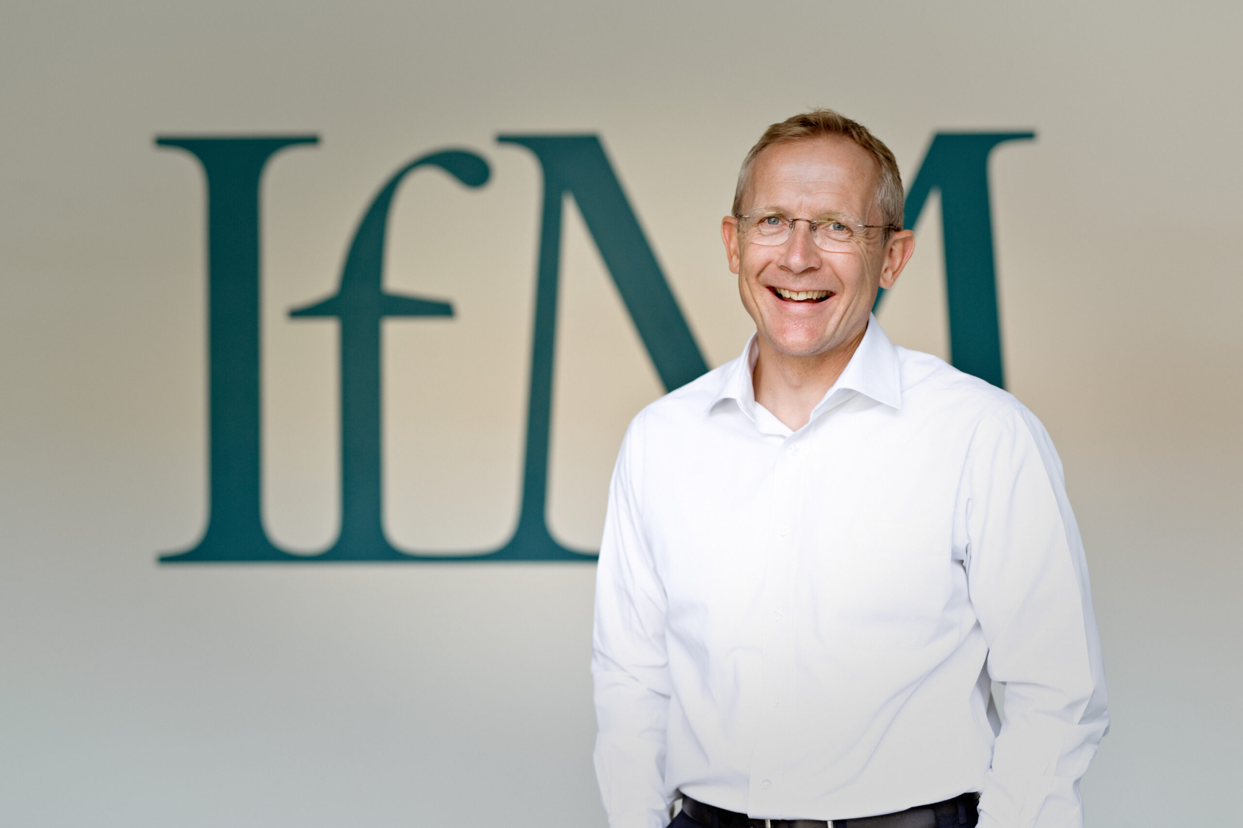 A smiling man wearing glasses and a white shirt stands in front of a wall with large teal letters IfM in the background.
