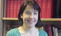 A woman is smiling in front of a bookshelf filled with red and black books. She has short dark hair and is wearing a green top.