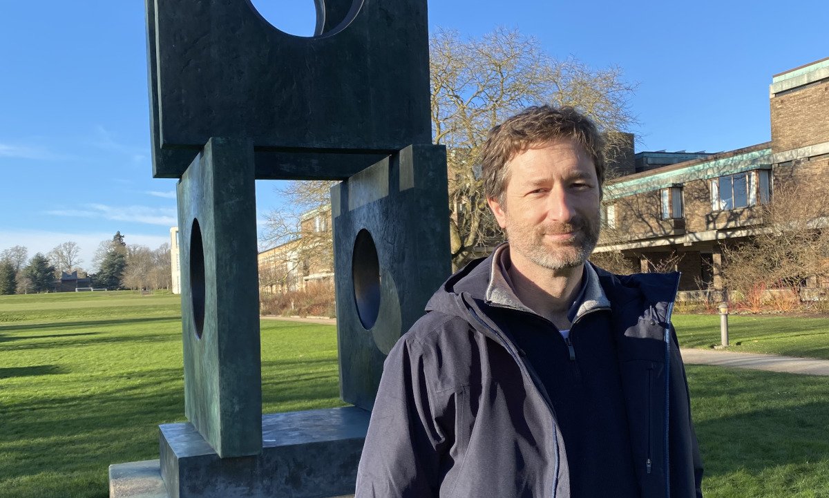 A person with short hair and a beard stands outdoors on a sunny day. He is wearing a dark jacket and standing in front of a modern sculpture with geometric shapes. The background features a green lawn and trees.