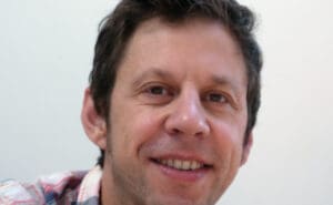 A person with short, dark hair smiling at the camera. They are wearing a plaid shirt. The background is plain and light-colored.