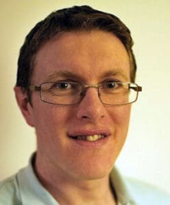 A person with short brown hair and glasses is smiling softly at the camera. They are wearing a light-colored shirt and are positioned against a plain background.