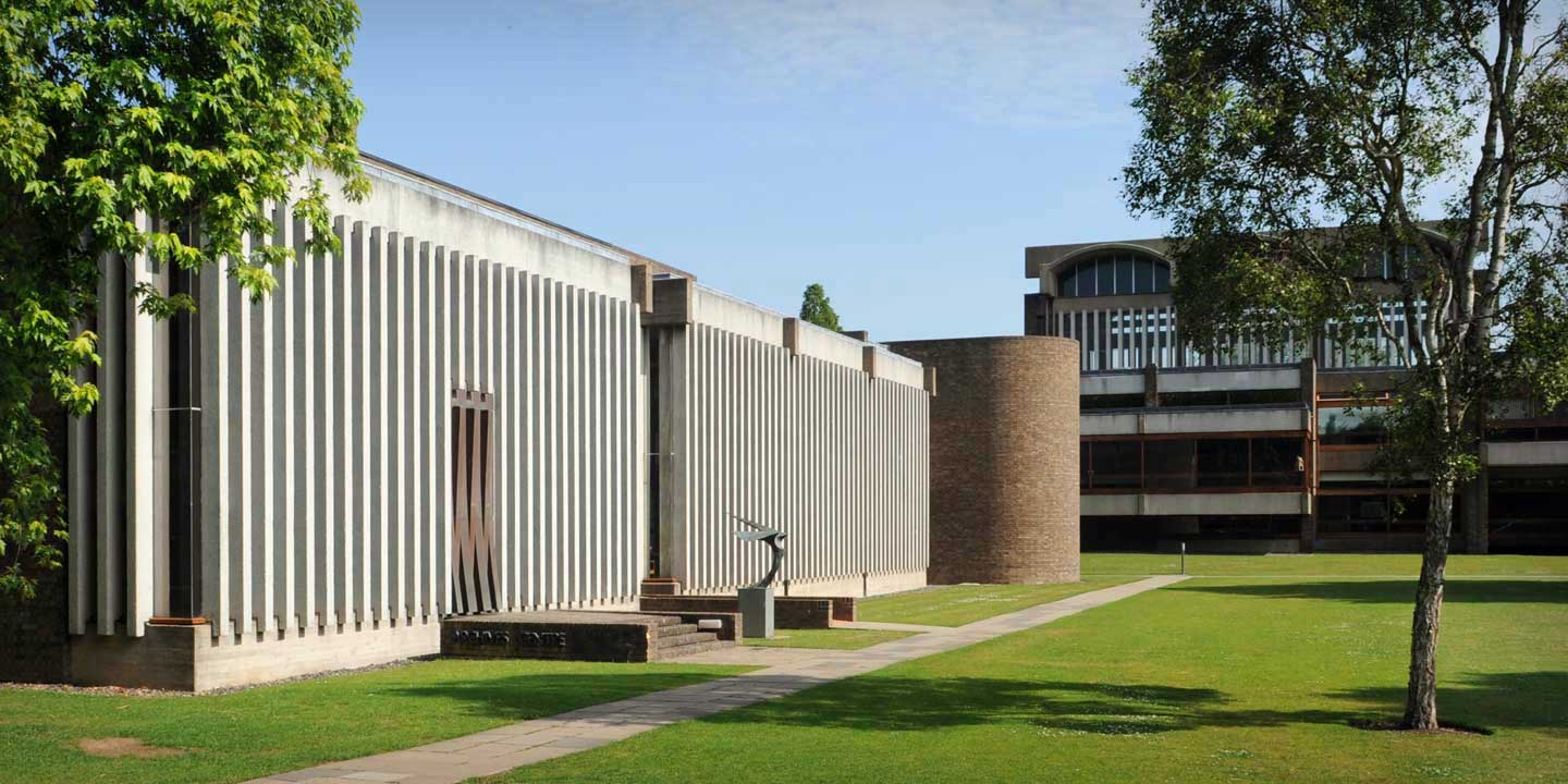A modern building with vertical concrete slats and a cylindrical section set in a lush green lawn. A sculpture is placed near the building on a small platform. Another structure with large windows is visible in the background.