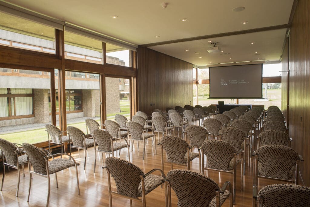 A room with rows of empty wicker chairs facing a projection screen. Large windows on the left side offer a view of a grassy area and a building outside. The room has a wooden floor and ceiling, creating a warm atmosphere.