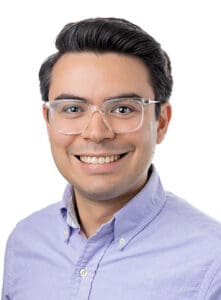 A person wearing glasses and a purple button-down shirt smiles at the camera against a white background.