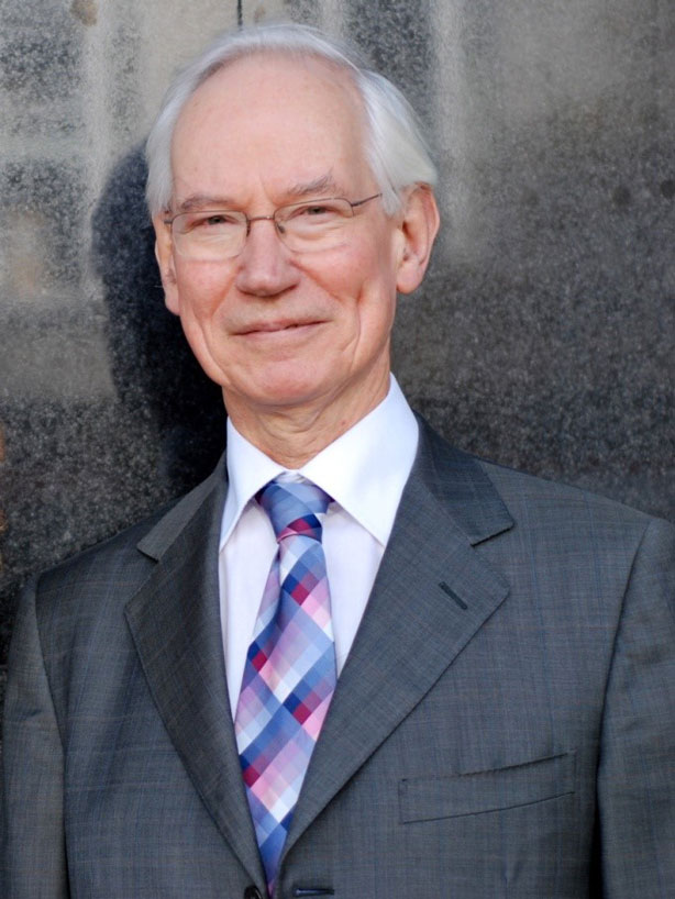 Man in a gray suit and checkered tie smiles slightly while standing against a textured gray wall.