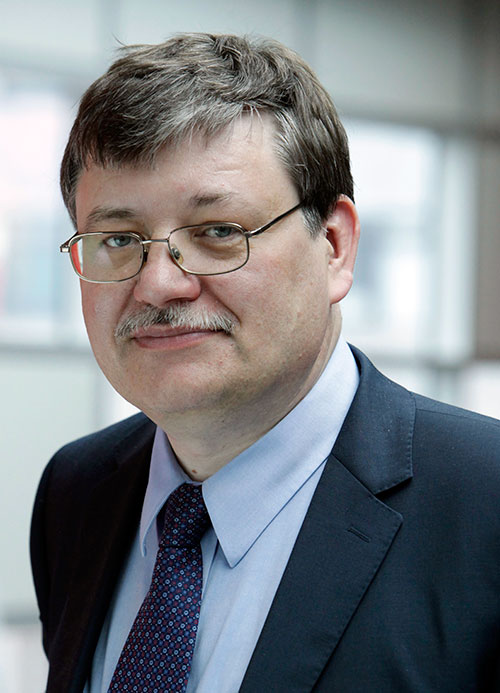 A man with glasses, short graying hair, and a mustache is wearing a suit with a light blue shirt and patterned tie. He is smiling slightly and standing in front of a blurred indoor background.