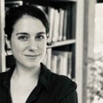 A person with dark hair is smiling slightly while standing in front of a bookshelf filled with books. The image is in black and white, giving it a classic look. There's a window in the background showing some natural light.
