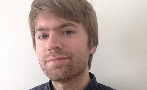 A person with short brown hair and light facial hair is wearing a dark shirt and looking at the camera with a neutral expression. The background is a plain white wall.