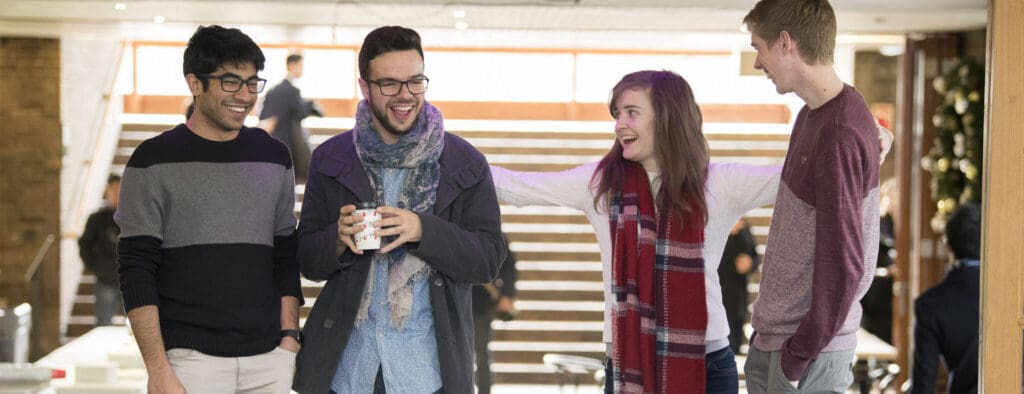 Four young adults, three men and one woman, stand indoors laughing and talking. Two hold mugs. They appear to be in a warmly lit communal space with stairs in the background.