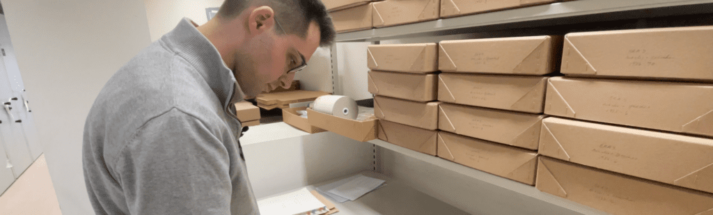 A person stands in a room, looking at documents. He wears glasses and a gray top. To the right, there are shelves filled with brown cardboard storage boxes. Papers and a roll of labels are on a desk.