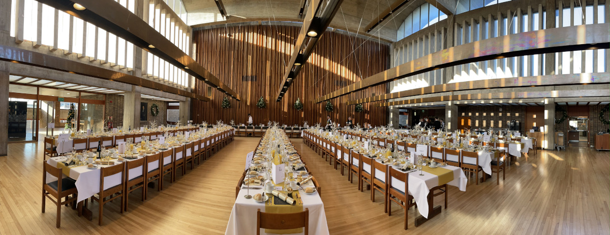 A large dining hall with long, parallel tables set for a formal event. Tables are adorned with white tablecloths, glasses, plates, and cutlery. Wooden panels and high windows add to the spacious ambiance.
