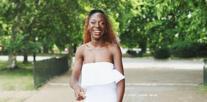 A smiling woman in a white strapless dress walks along a tree-lined path, with sunlight filtering through the leaves. The path is surrounded by lush greenery, creating a vibrant, natural backdrop.