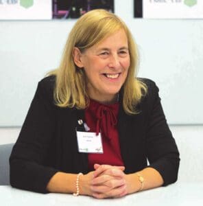 A woman with blonde hair sits at a table, smiling. She is wearing a black blazer over a red blouse and has a name tag. Her hands are clasped in front of her, and she wears a pearl bracelet. The background shows part of a wall and a framed picture.