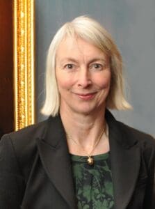 A woman with shoulder-length blonde hair wearing a black blazer and a dark green patterned blouse, smiles at the camera. The background shows part of a gold-framed picture against a gray wall.