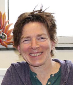 A person with short, tousled hair smiles warmly at the camera. They are wearing a casual shirt and hoodie. A colorful, decorative object is partially visible in the background next to a window.