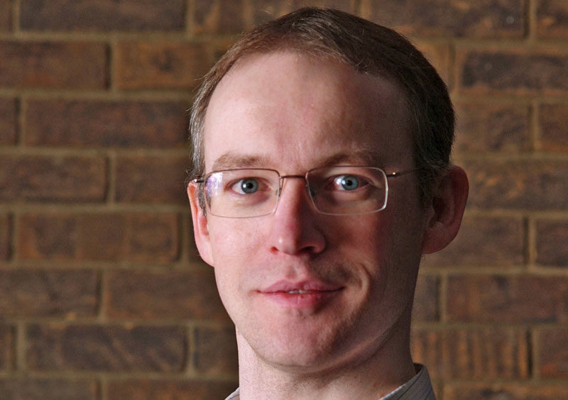 A person with light skin and short, light brown hair is wearing glasses and a collared shirt, standing in front of a brick wall. The person is smiling slightly and looking at the camera.