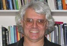 A man with gray hair and glasses smiling in front of a bookshelf filled with various books.