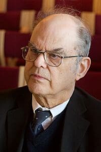 A man with glasses and a serious expression is wearing a black suit, dark sweater, white shirt, and patterned tie. He is indoors, against a blurred background of maroon and beige seats.