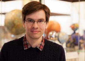 A person with short brown hair and glasses is smiling, wearing a dark sweater and a red plaid shirt. He is standing in front of a display with various globes and spherical objects, slightly out of focus.