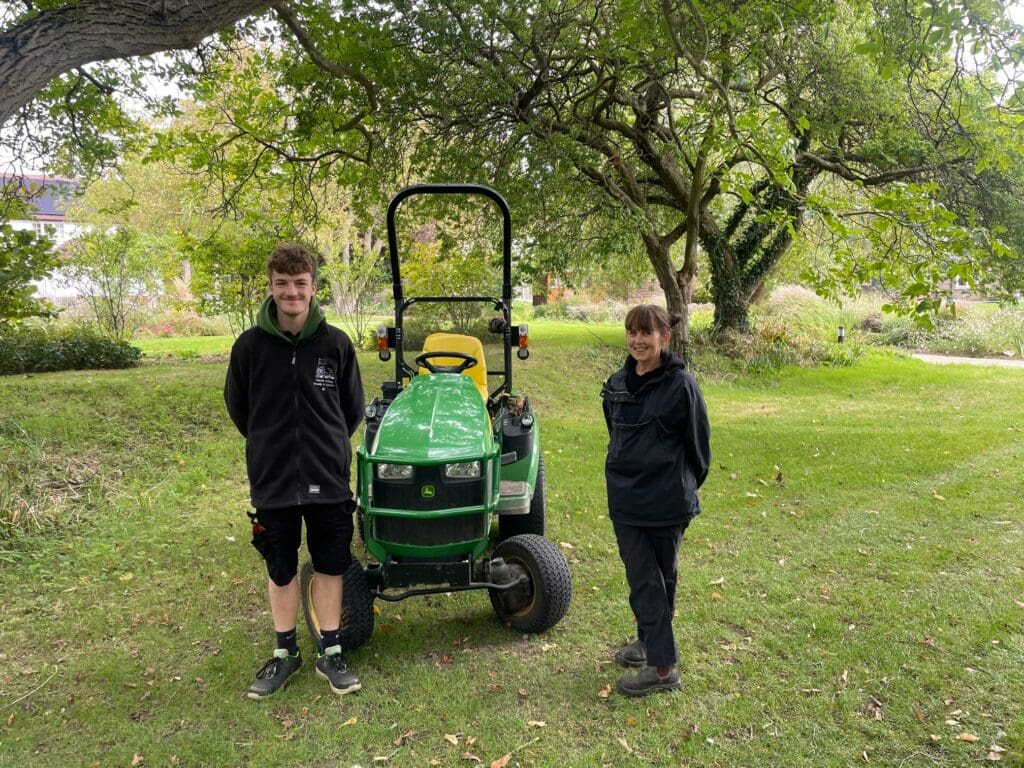Two people in dark jackets stand on a grassy area in a park. A green ride-on lawnmower is between them. Trees and shrubs surround the scene, with a path visible in the background.