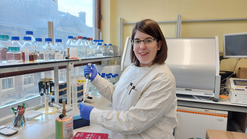 A person in a lab coat and gloves uses a pipette at a laboratory bench. Shelves behind her hold various bottles. A computer and lab equipment are visible nearby. A window shows a building outside.