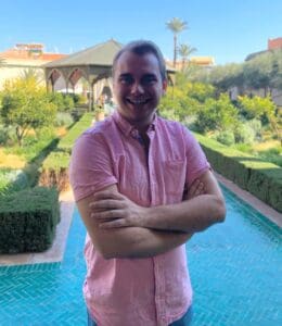 A person in a pink shirt stands with arms crossed, smiling in front of a turquoise-tiled path. The background features a garden with symmetrical hedges, bushes, and palm trees under a clear blue sky.