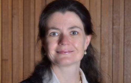 A person with long dark hair, wearing a white collared shirt and a necklace, stands against a wooden-paneled background. They are smiling slightly and looking directly at the camera.
