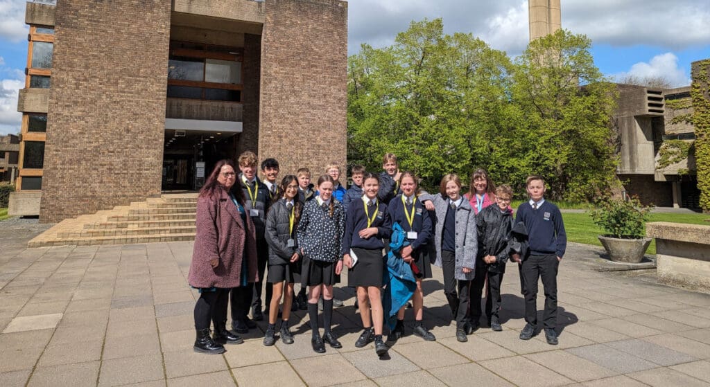 A group of people, including teenagers and adults, stand smiling on a paved area in front of a modern brick building. Some wear school uniforms, and the background includes trees and a cloudy sky.