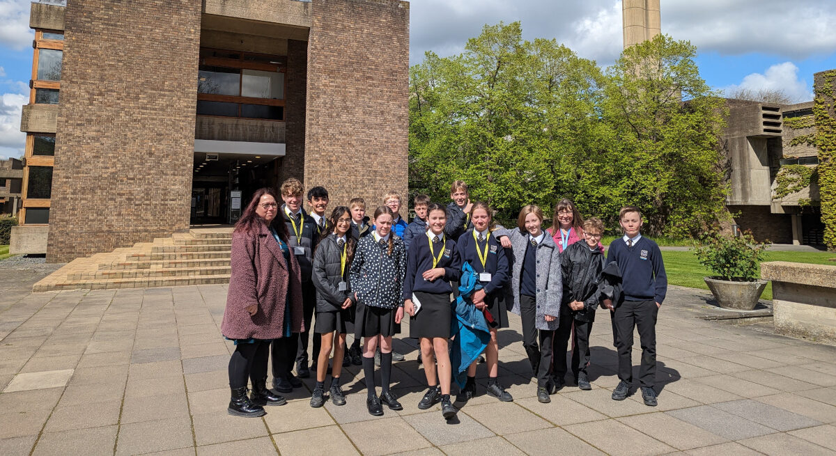 A group of people, including teenagers and adults, stand smiling on a paved area in front of a modern brick building. Some wear school uniforms, and the background includes trees and a cloudy sky.