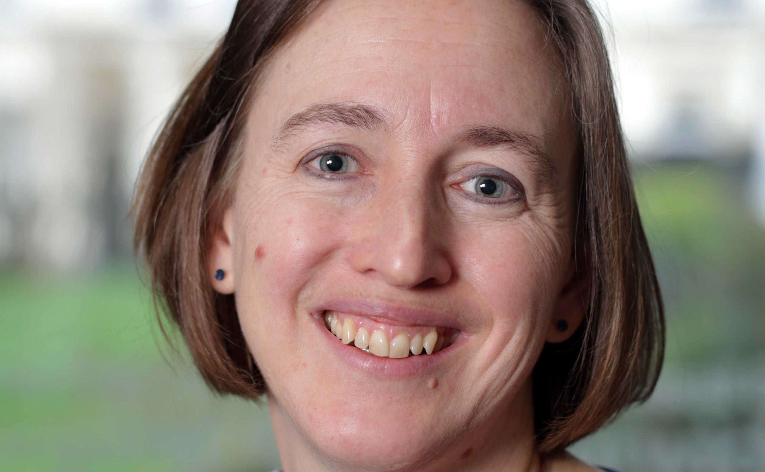 A person with short brown hair smiles at the camera, wearing a light-colored top and small stud earrings. The background is softly blurred green and white tones.