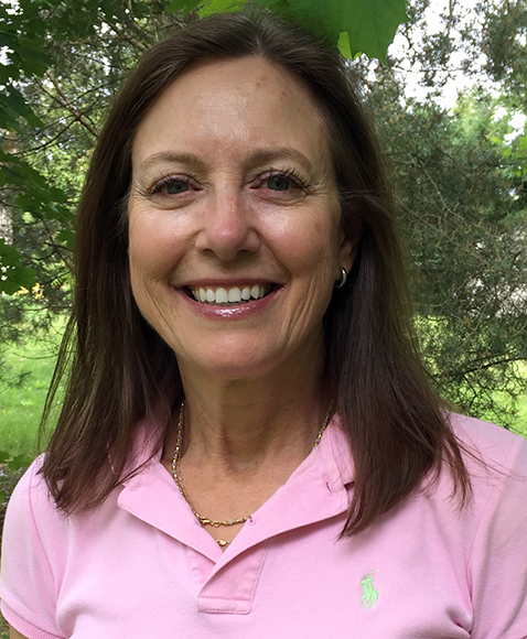 A woman with long brown hair smiles while wearing a pink collared shirt with a small green logo. She stands outdoors, surrounded by greenery, with trees and grass in the background.