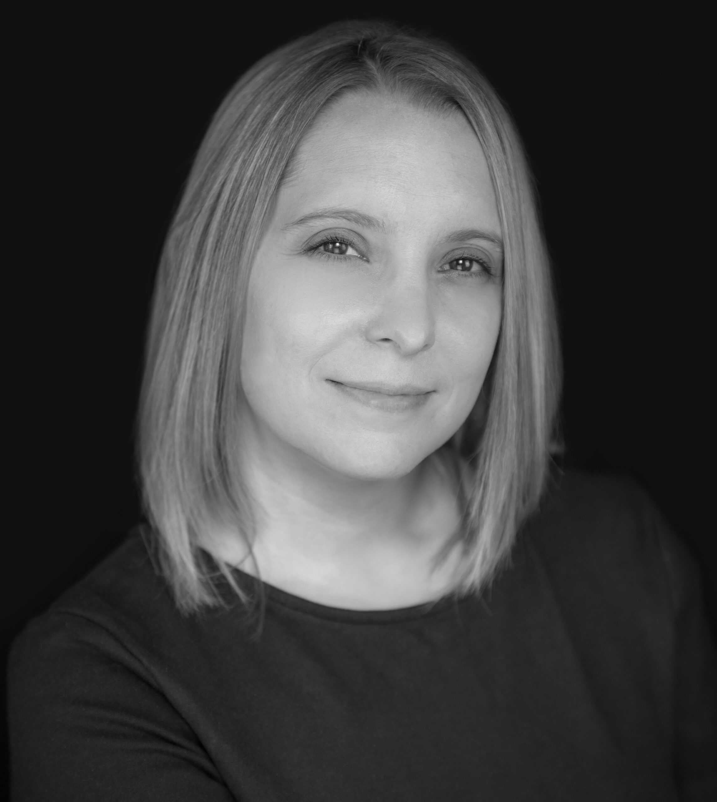 A black and white portrait of a person with shoulder-length blonde hair, wearing a dark top, smiling softly at the camera against a dark background.